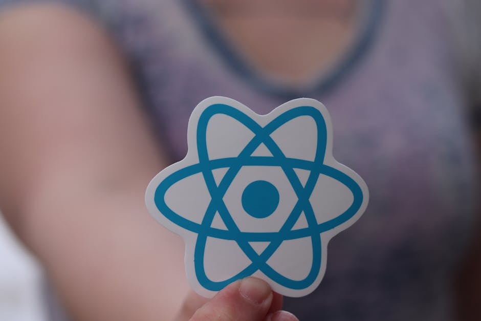 Close-up of a person's hand holding a sticker featuring a blue atomic model symbol, with the person's arm and torso visible in the background. The background includes a blurred view of a room with neu