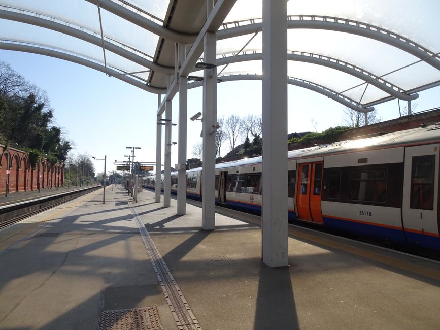 View of a modern, well-maintained railway station platform through an oval-shaped window, featuring clean wooden wall panels, a smooth concrete floor, and neatly arranged turnstiles with stainless ste